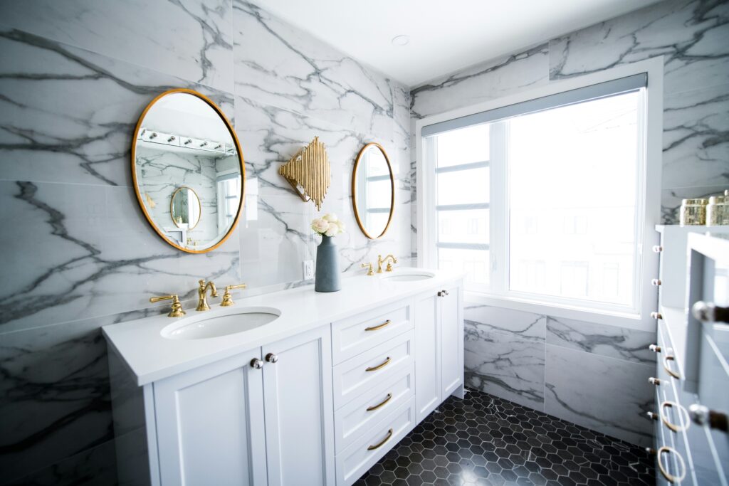Luxurious bathroom with marble walls, dual sinks with gold fixtures, oval mirrors, and a hexagon-tiled floor.