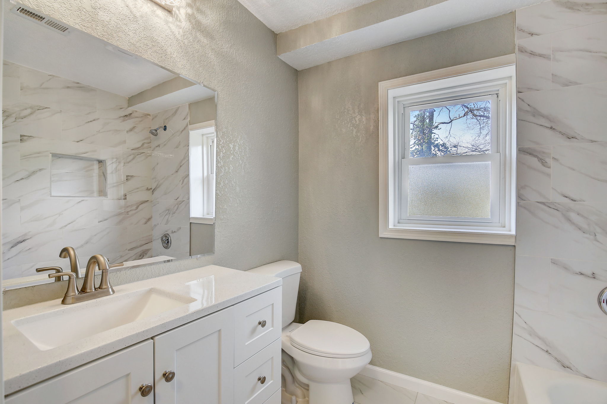 A modern bathroom featuring a white vanity with a quartz countertop, a brushed nickel faucet, a toilet, and a bathtub with white marble tile surround and a recessed shelf.